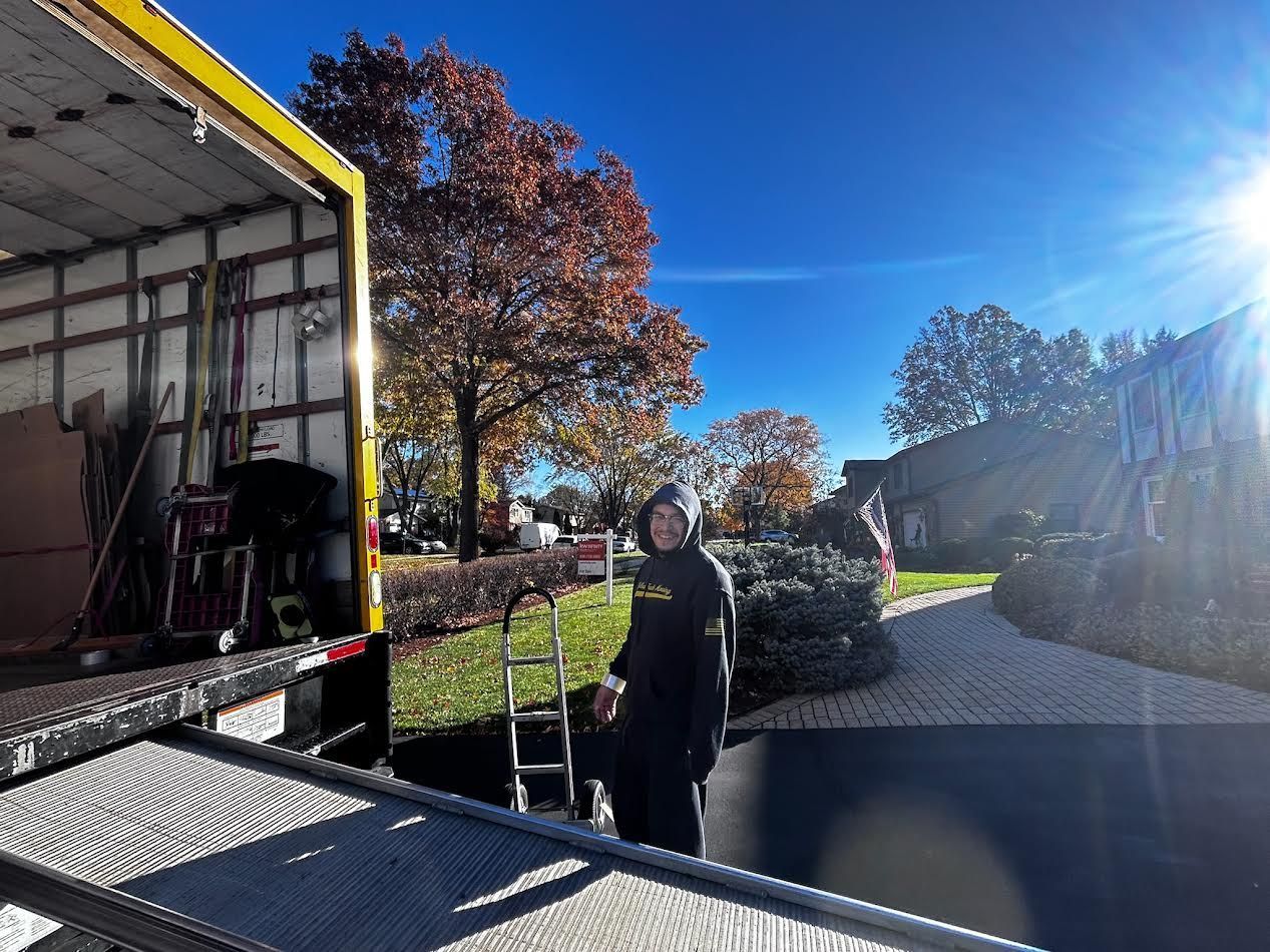Person standing by the open back of a moving truck on a sunny day with fall foliage.