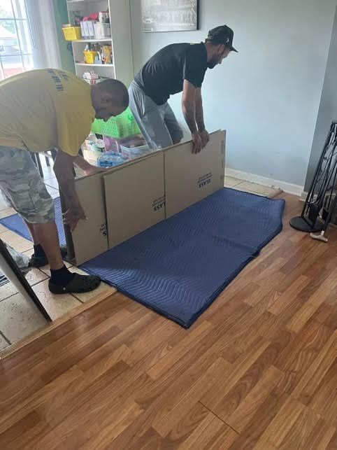 Two people lift a large cardboard box over a blue moving blanket inside a home.