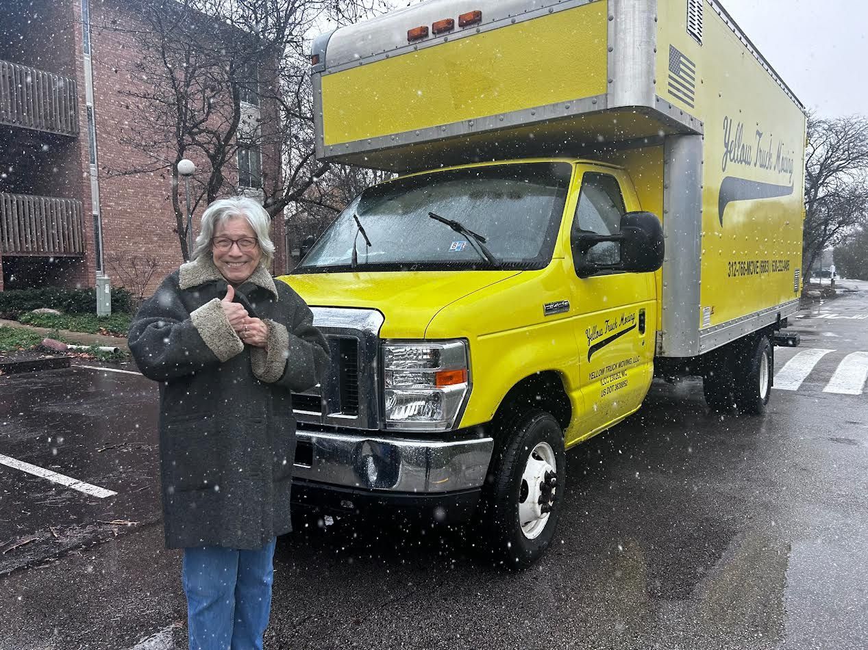 Man in coat giving thumbs up next to a bright yellow moving truck in a snowy street.