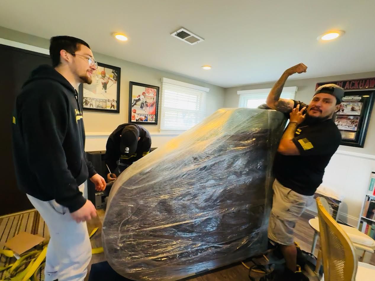 Movers wrapping furniture in plastic. Two men in a room; one flexes his arm.