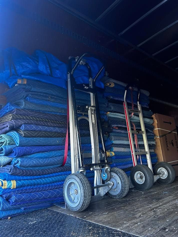 Inside a truck, two hand trucks stand next to stacked blue moving blankets.