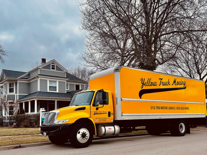Yellow moving truck parked in front of a gray house on a cloudy day.