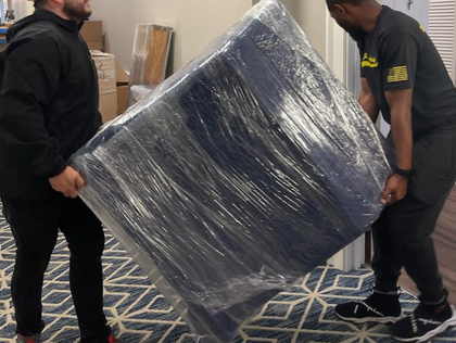 Two people carrying a large, blue, plastic-wrapped object through a hallway.