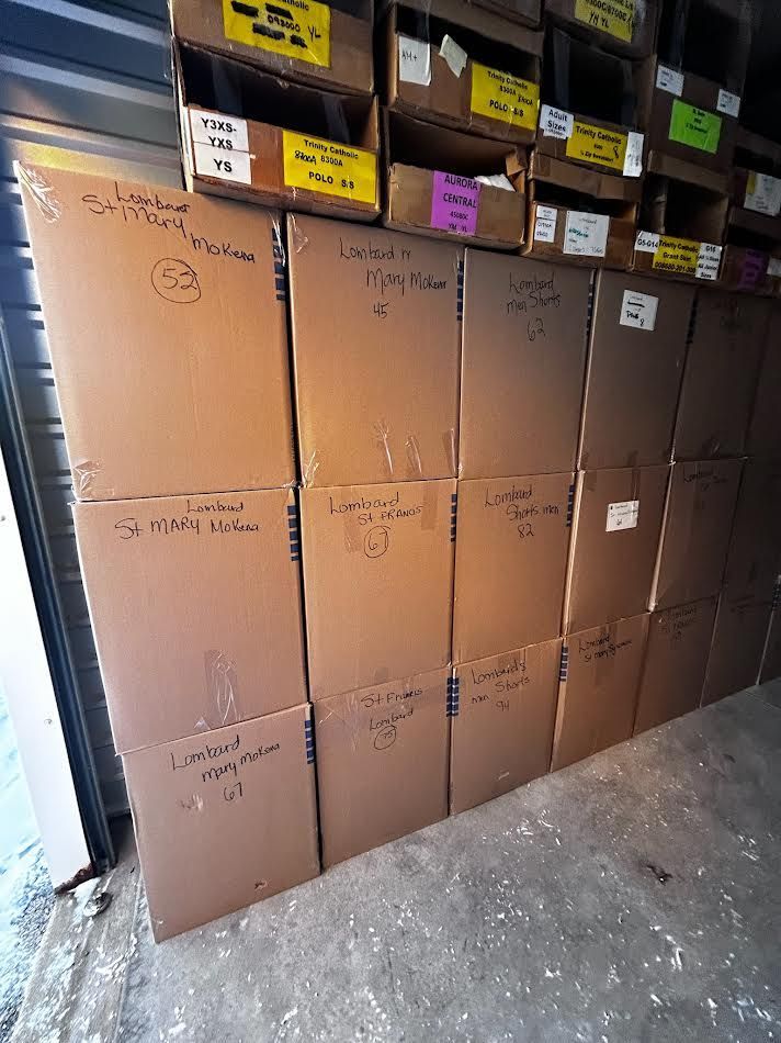 Cardboard boxes stacked in a storage unit, labeled with handwritten text. More boxes on shelves above.