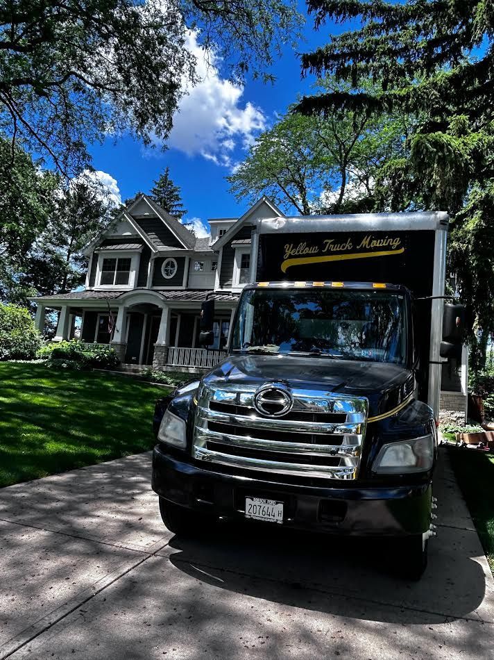 Moving truck parked in front of a large house with a green lawn and trees under a blue sky.