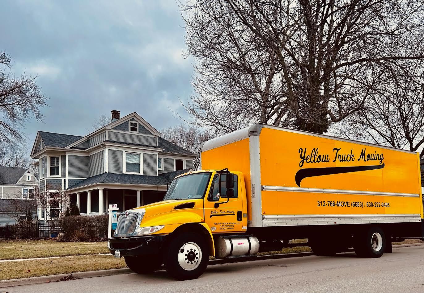 Yellow moving truck parked on a residential street in front of a gray house on a cloudy day.