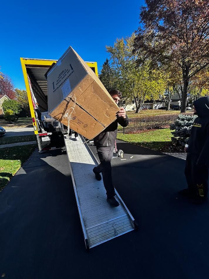 Man carrying large cardboard box down a ramp from a moving truck on a sunny day.