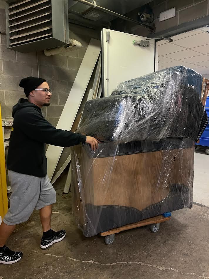 Man pushing a large, wrapped furniture item on a dolly in an industrial setting.