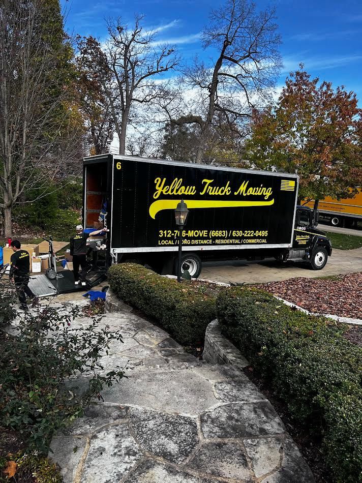 Yellow Track Moving truck parked on a driveway with workers loading boxes. Autumn foliage in background.