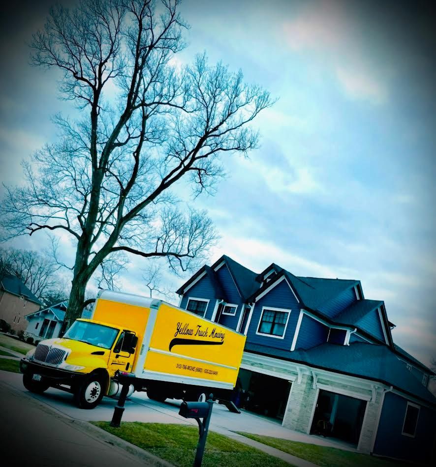 Yellow moving truck parked in front of a blue house with open garage doors. Tree and cloudy sky in background.