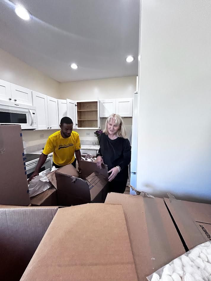 Two people unpacking boxes in a new kitchen with white cabinets. One person wears a yellow shirt.