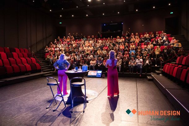 Twee vrouwen staan ​​op het podium voor een groot publiek in een theater. De ene vrouw draagt ​​een roze jurk, de andere een rode.