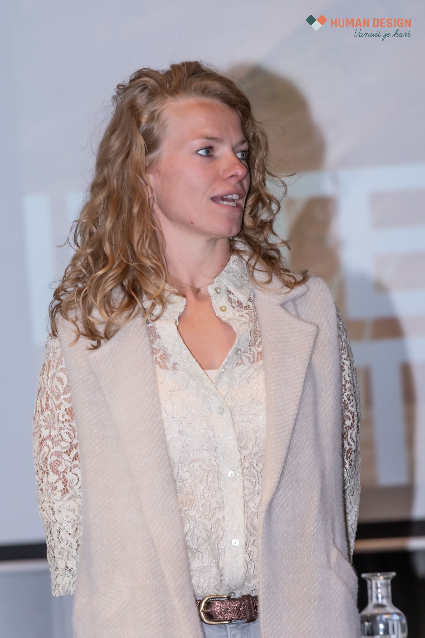 Vrouw in paarse jumpsuit, met bril, staat achter een podium en spreekt. Presentatie op de achtergrond.