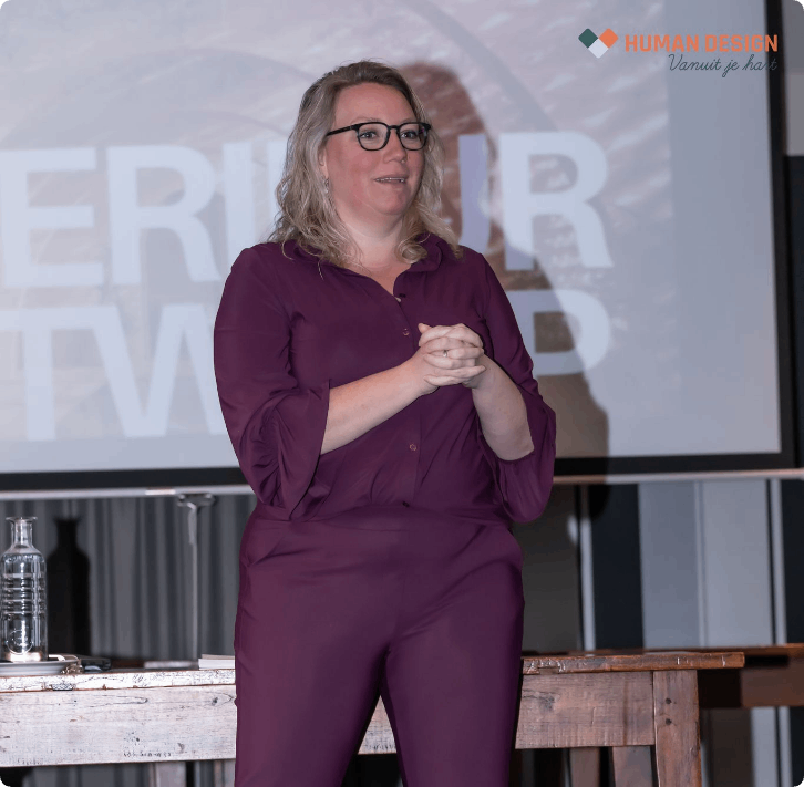 Vrouw in paarse jumpsuit, met bril, staat achter een podium en spreekt. Presentatie op de achtergrond.