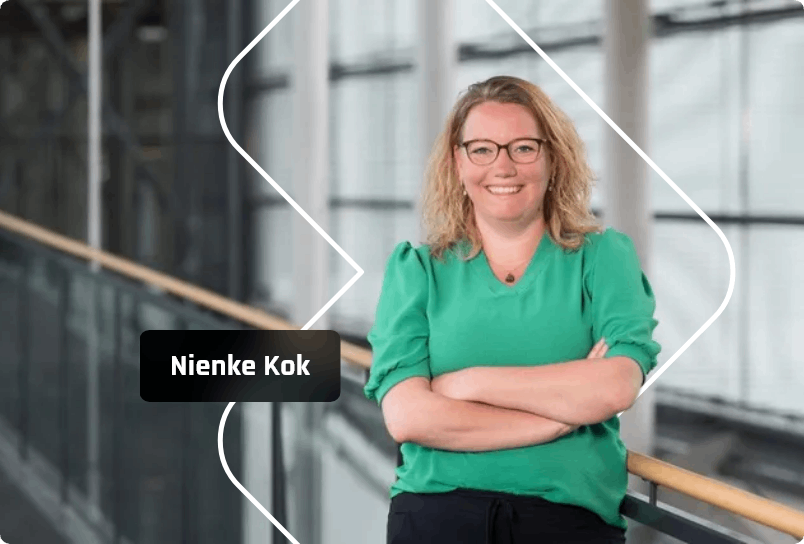Nienke Kok, een lachende vrouw met gekruiste armen, een bril en een groene top, staat binnen met een architectonische achtergrond.
