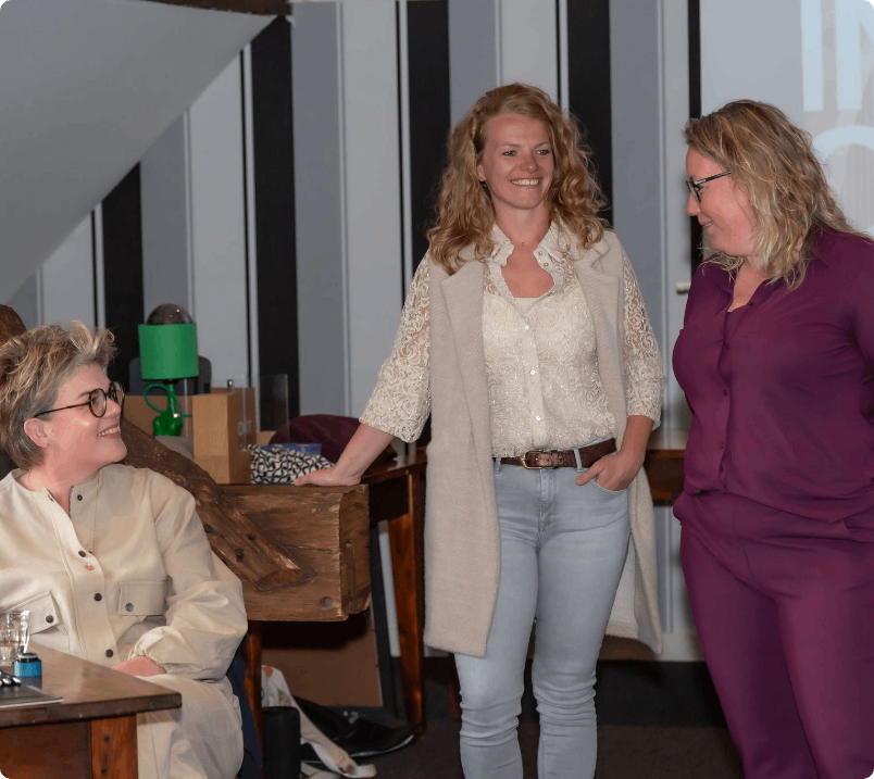 Drie vrouwen in een kamer, twee staan, één zit. Ze glimlachen en zijn in gesprek. Neutrale achtergrond, gedempt licht.