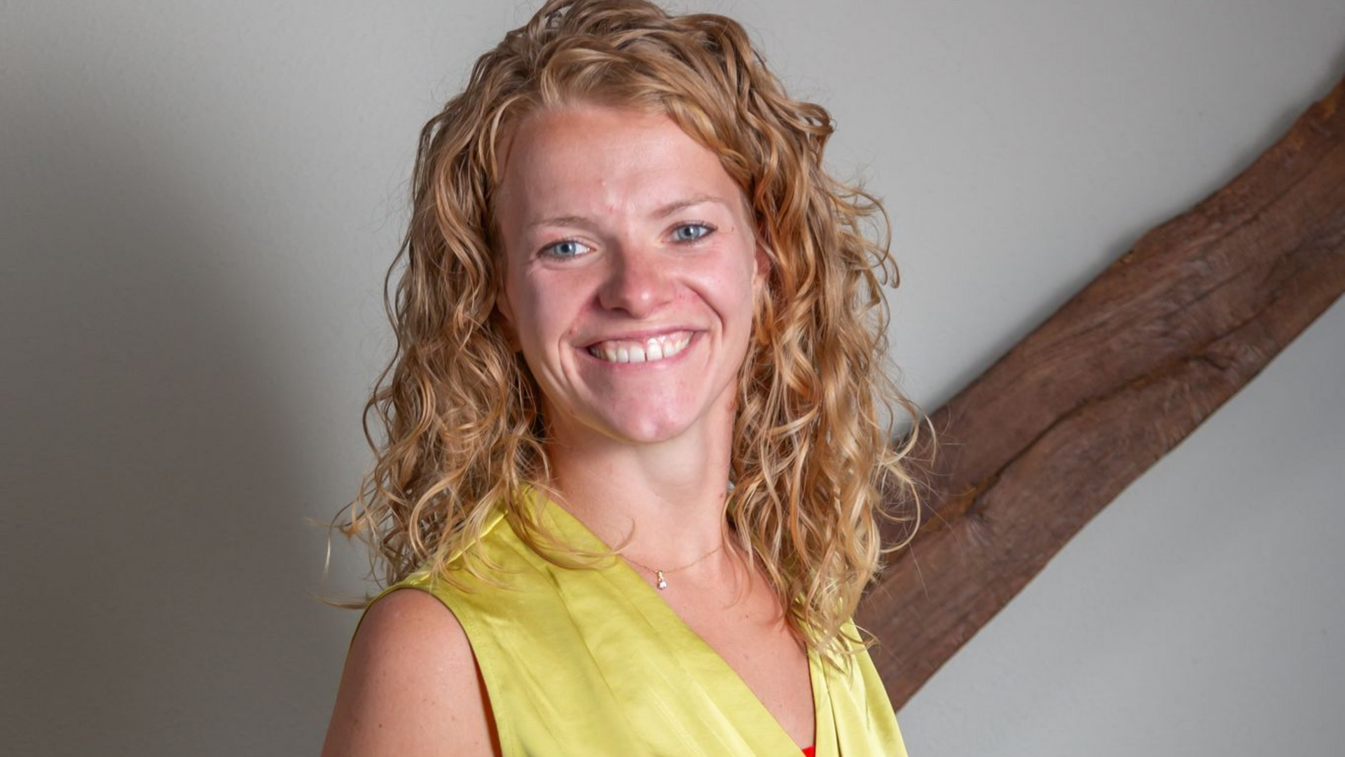 Nienke Kok, een lachende vrouw met gekruiste armen, een bril en een groene top, staat binnen met een architectonische achtergrond.