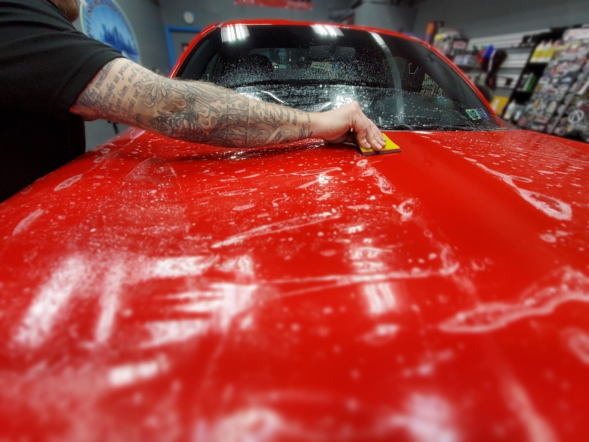 A man is applying a protective film to the hood of a red car.