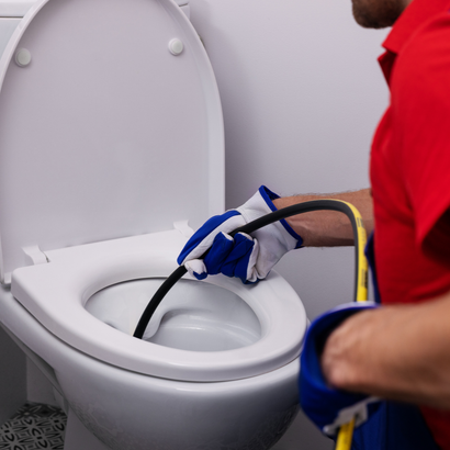 Person in red shirt and gloves uses a plumbing snake to unclog a white toilet.