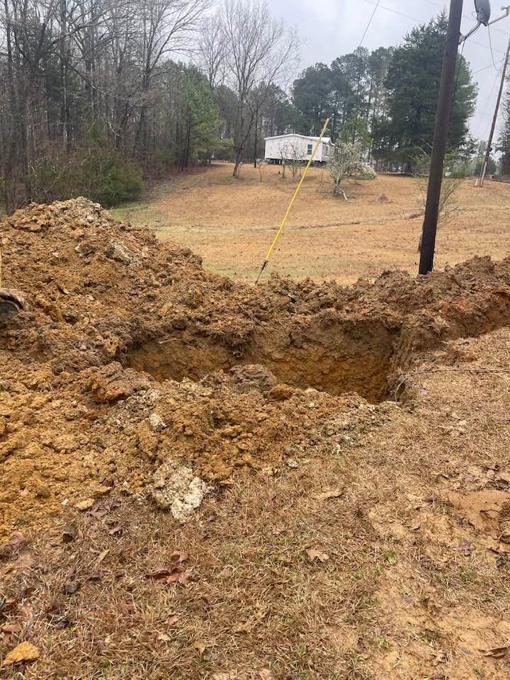 A large hole dug in brown earth, with a utility pole and a mobile home visible in the background.