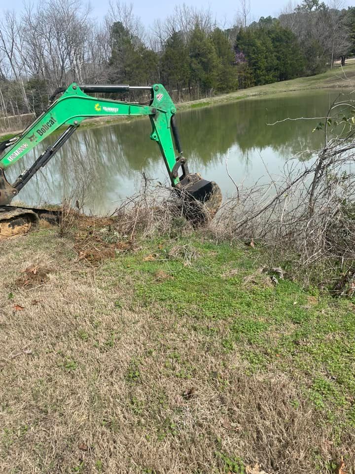 A green excavator working on the edge of a pond, clearing brush and dirt.