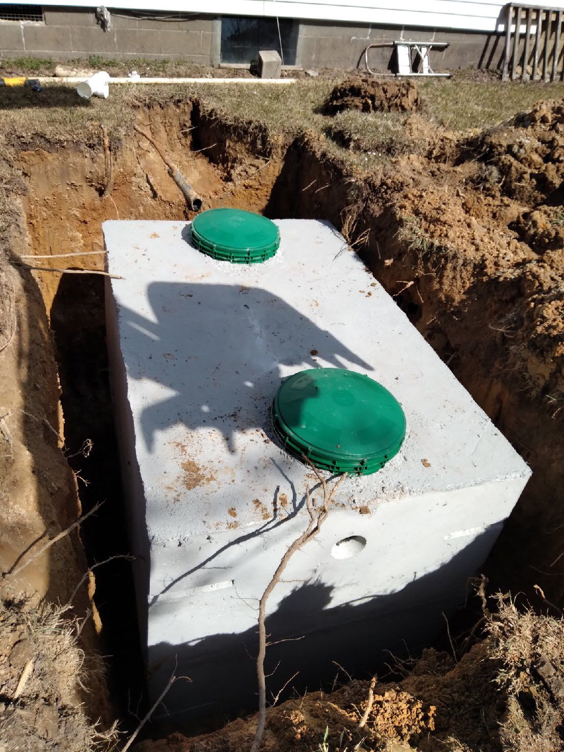Concrete septic tank with two green lids in an excavated pit.