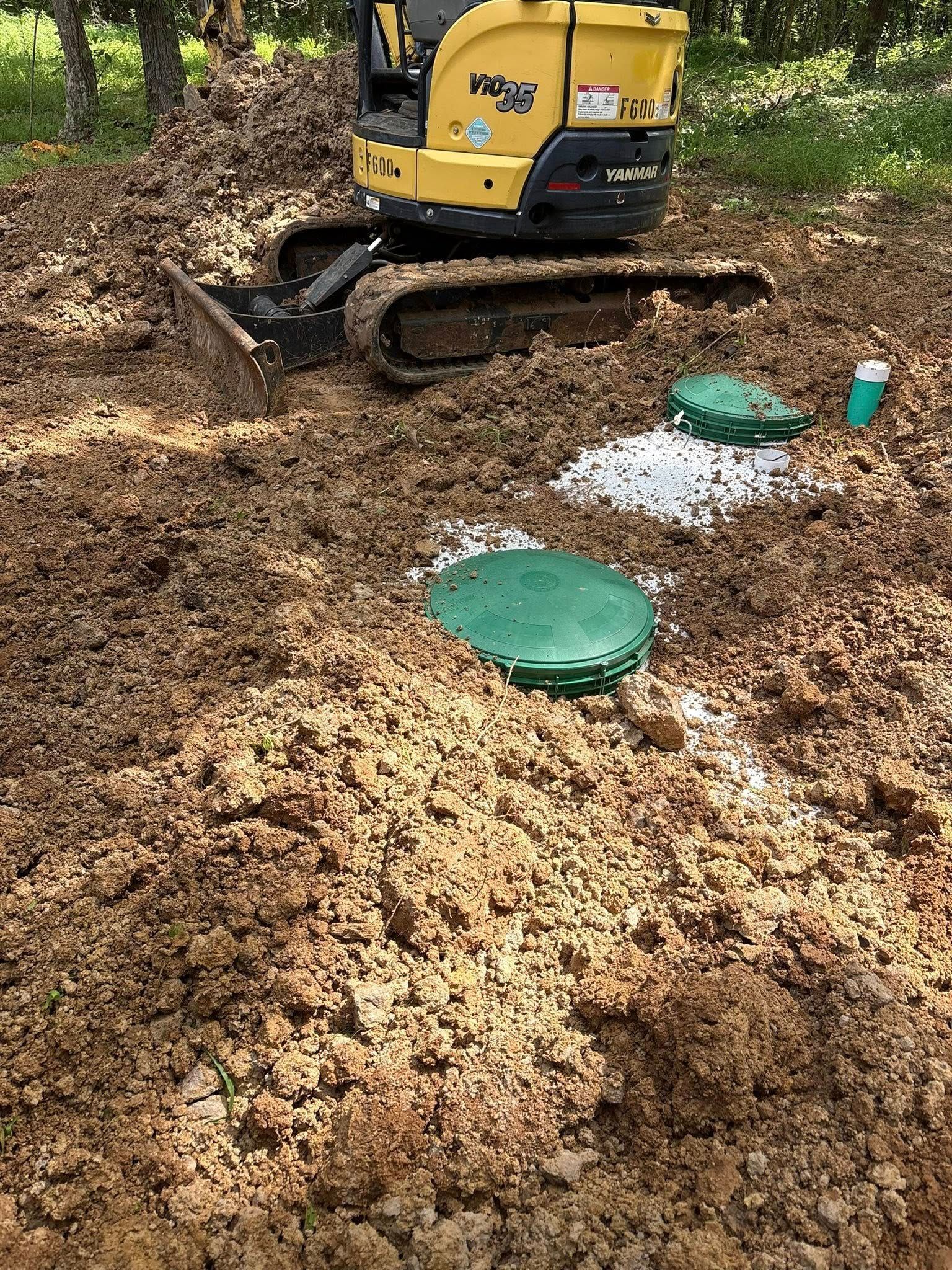 Yellow excavator over dirt. Green septic tank lids are visible.
