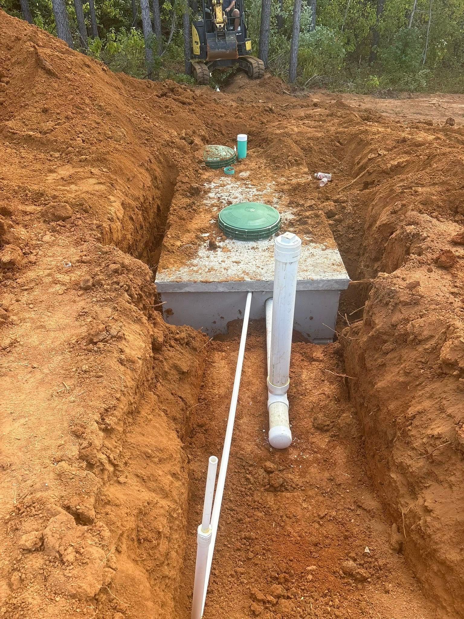 Septic tank installation in red soil, with white PVC pipes and a green lid.