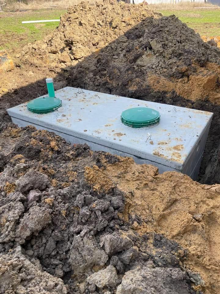 A rectangular concrete septic tank partially buried in a dirt trench, with two green access covers and a vent pipe.