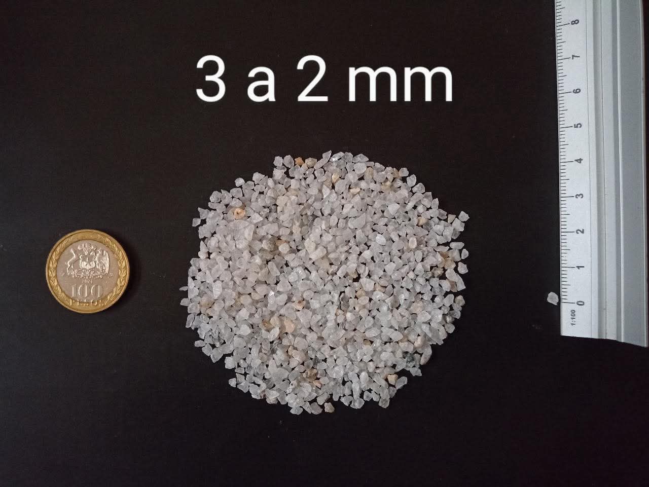 Montón de grava de color claro, de 2 a 3 mm de tamaño, junto a una moneda y una regla sobre una superficie negra.
