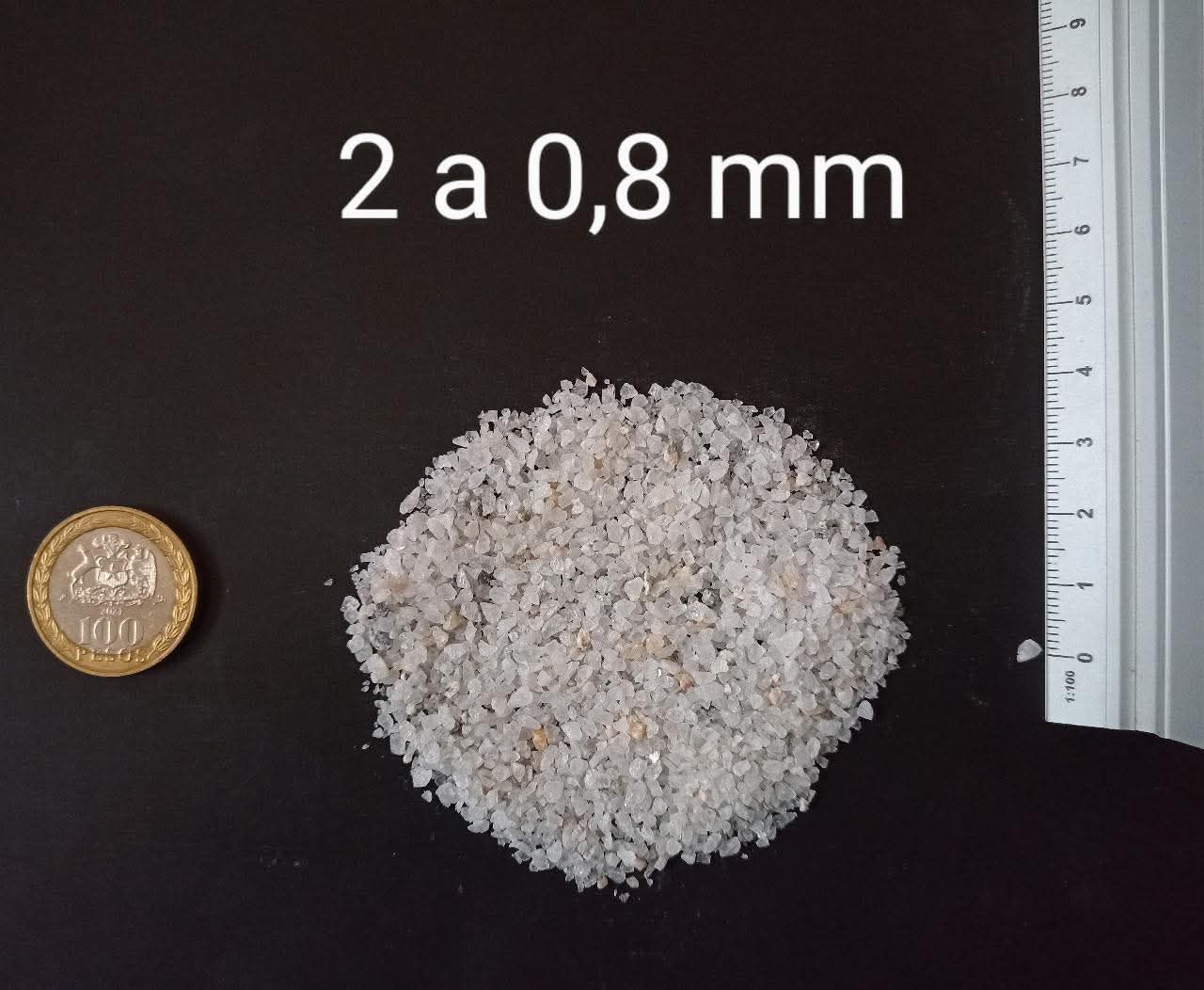 Montón de material parecido a la arena con una moneda como referencia de tamaño, junto a una regla; el tamaño se especifica entre 2 y 0,8 mm.