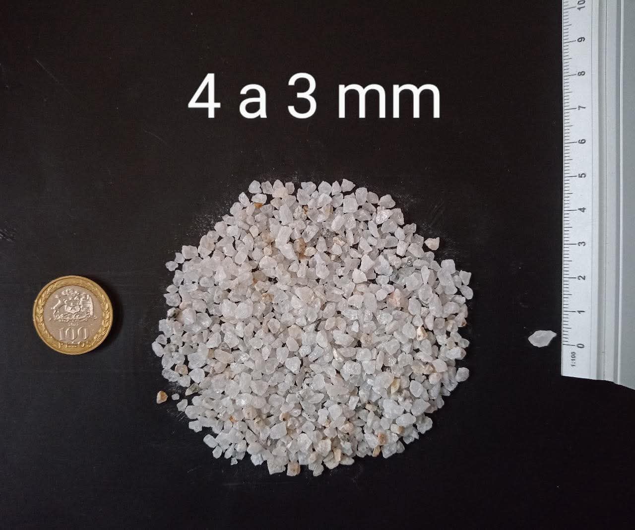 Montón de pequeñas piedras de color claro, de 3 a 4 mm de tamaño, junto a una moneda y una regla sobre una superficie oscura.