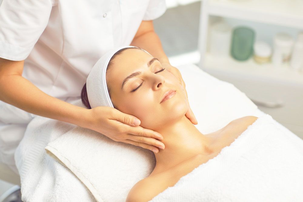 Woman receiving a facial massage at a spa. She is relaxed with eyes closed, head wrapped in a white headband.