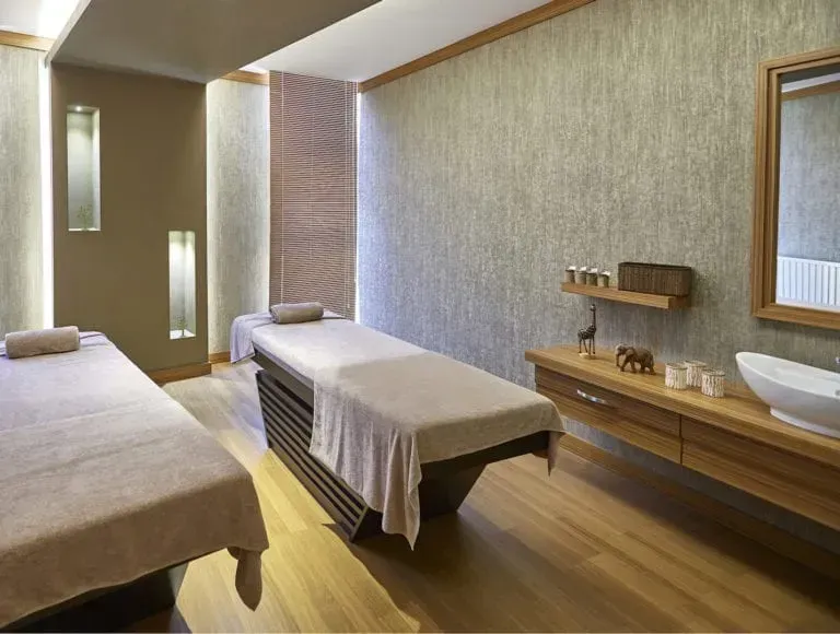 Two massage tables in a spa room with light brown walls, wood accents, and a sink area.
