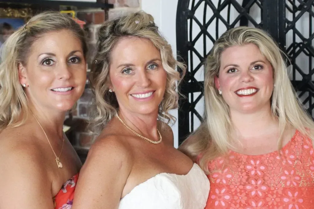 Three women smiling, one in a white wedding dress, blonde hair, outdoors, sunny day.