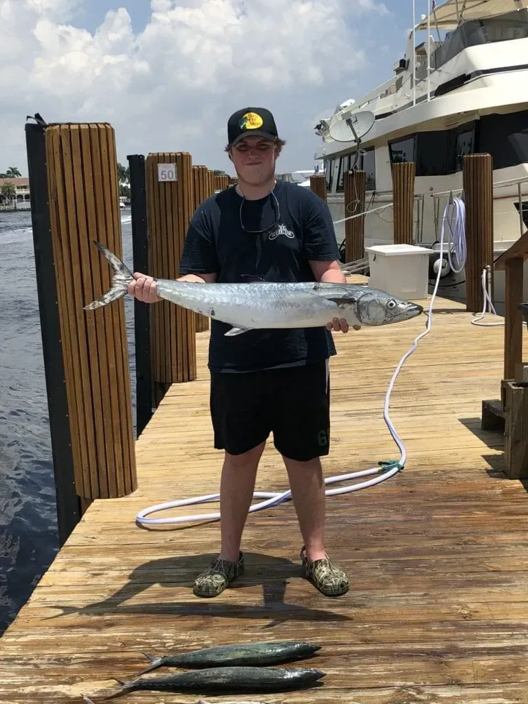 Teenager holding a fish on a dock. Two other fish are on the dock. Boat in the background.