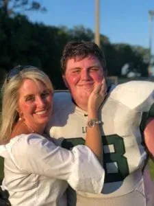 Woman in white shirt gently touches her son's cheek. The man wears a football jersey with green and white accents.