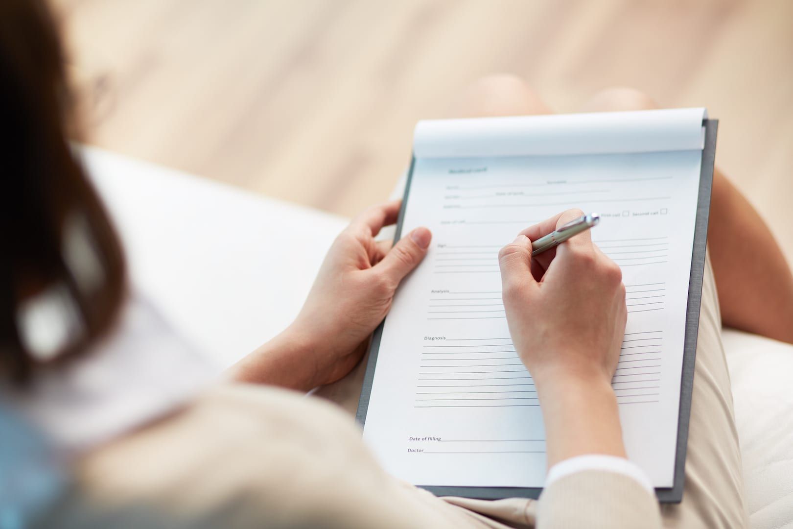 Person sitting, holding a clipboard with a form and writing with a pen. Beige suit, light background.