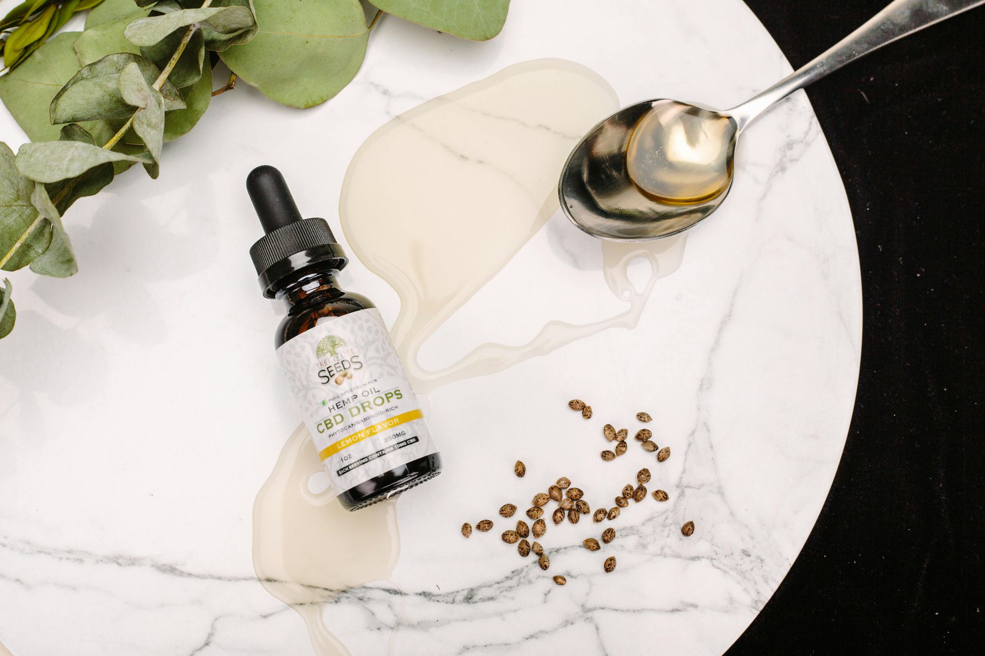 A bottle of CBD oil with a dropper sits on a marble surface, with a spoon, spilled liquid, and seeds, along with a branch.