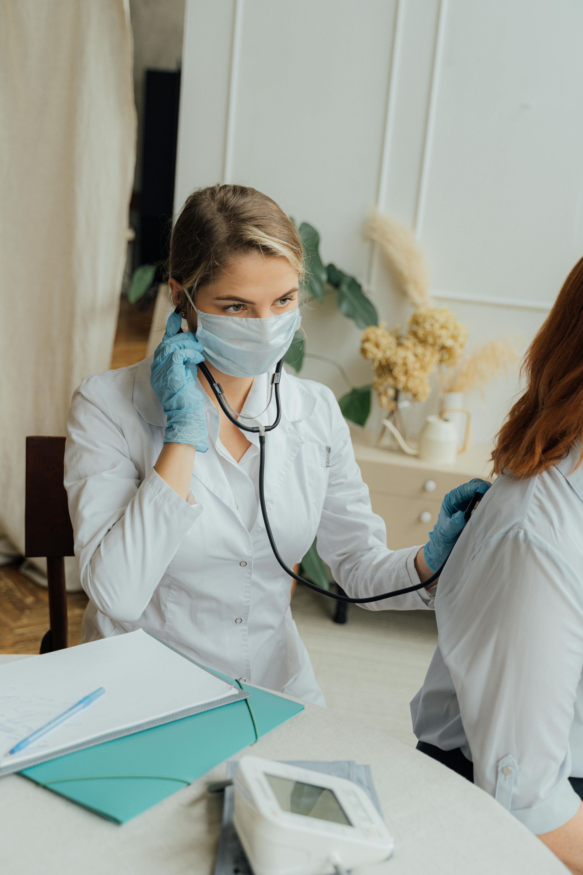 Doctor wearing a mask and gloves listens to a patient's chest with a stethoscope in a medical office setting.