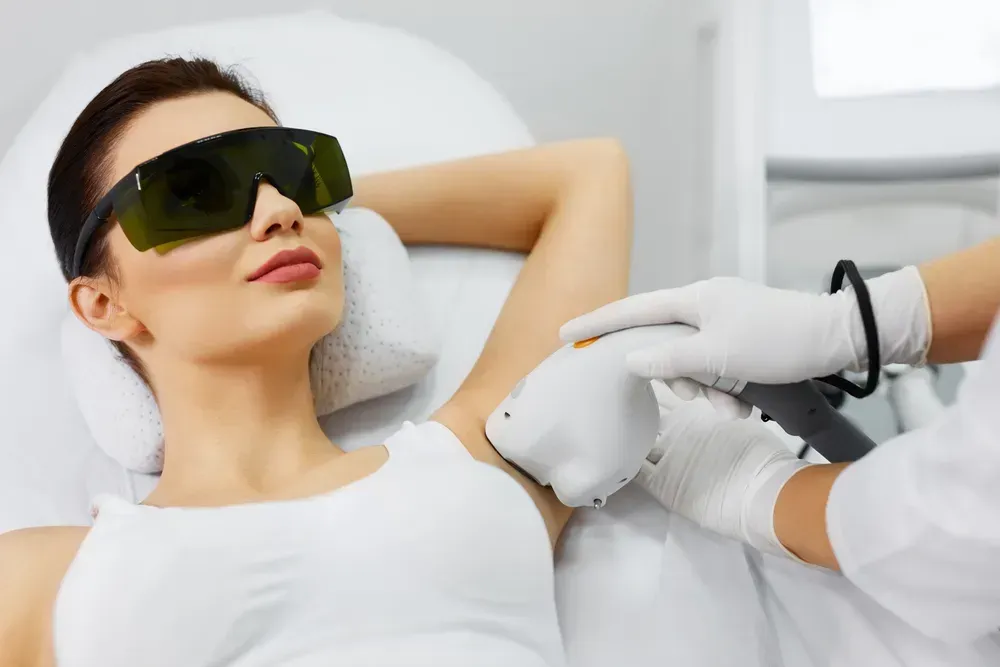 Woman receiving laser hair removal on her armpit while wearing protective eyewear. The medical setting is a spa or clinic.