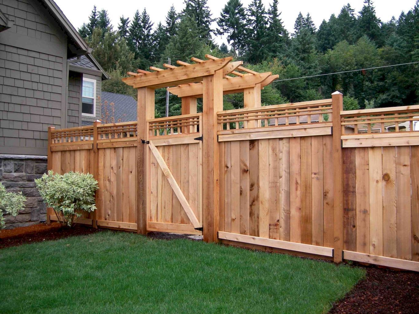 Wooden fence with gate and pergola over entrance; green lawn and trees in background.