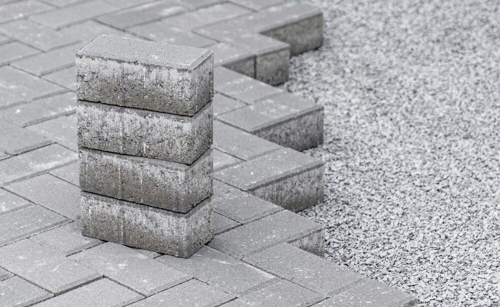 Stack of gray paving stones next to a partially laid gray brick patio on gravel.