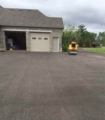 Asphalt driveway with garage; a yellow roller compacts fresh asphalt.