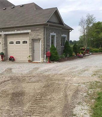 Gray stone building with garage, gravel driveway. Red mailbox, landscaping, cloudy sky.