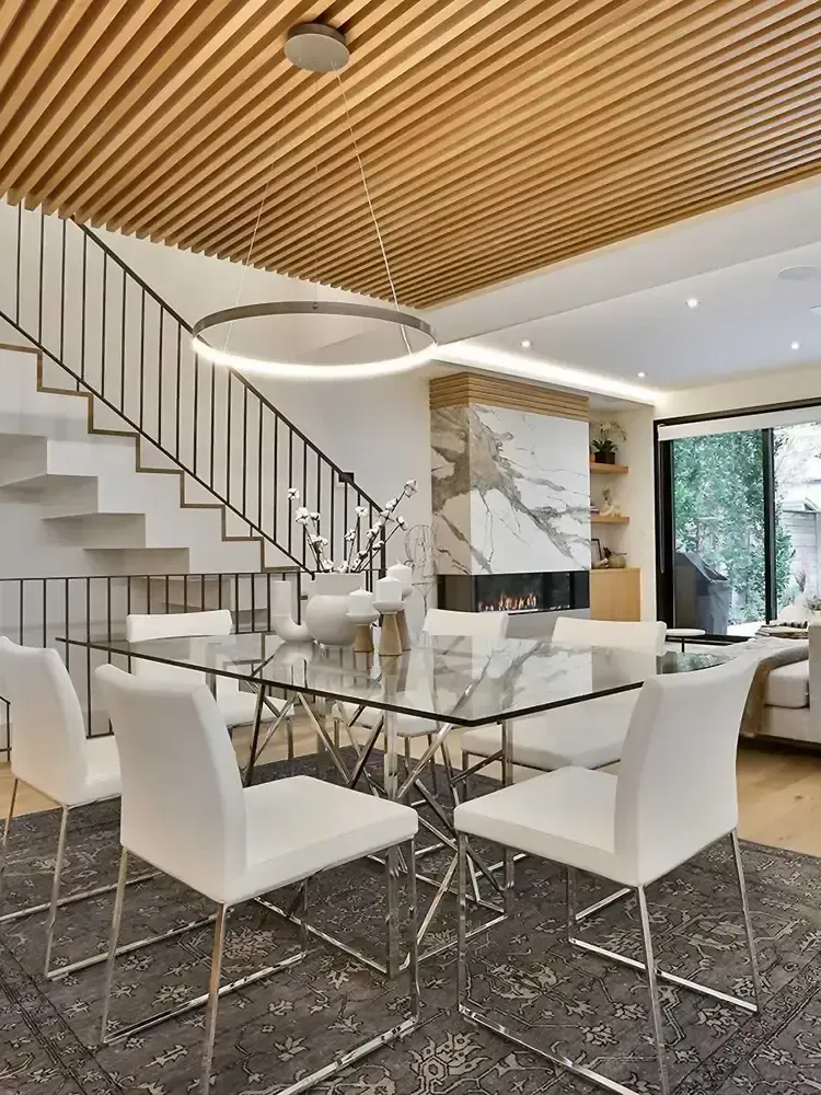 Modern dining room with glass table, white chairs, wooden ceiling, and marble fireplace.