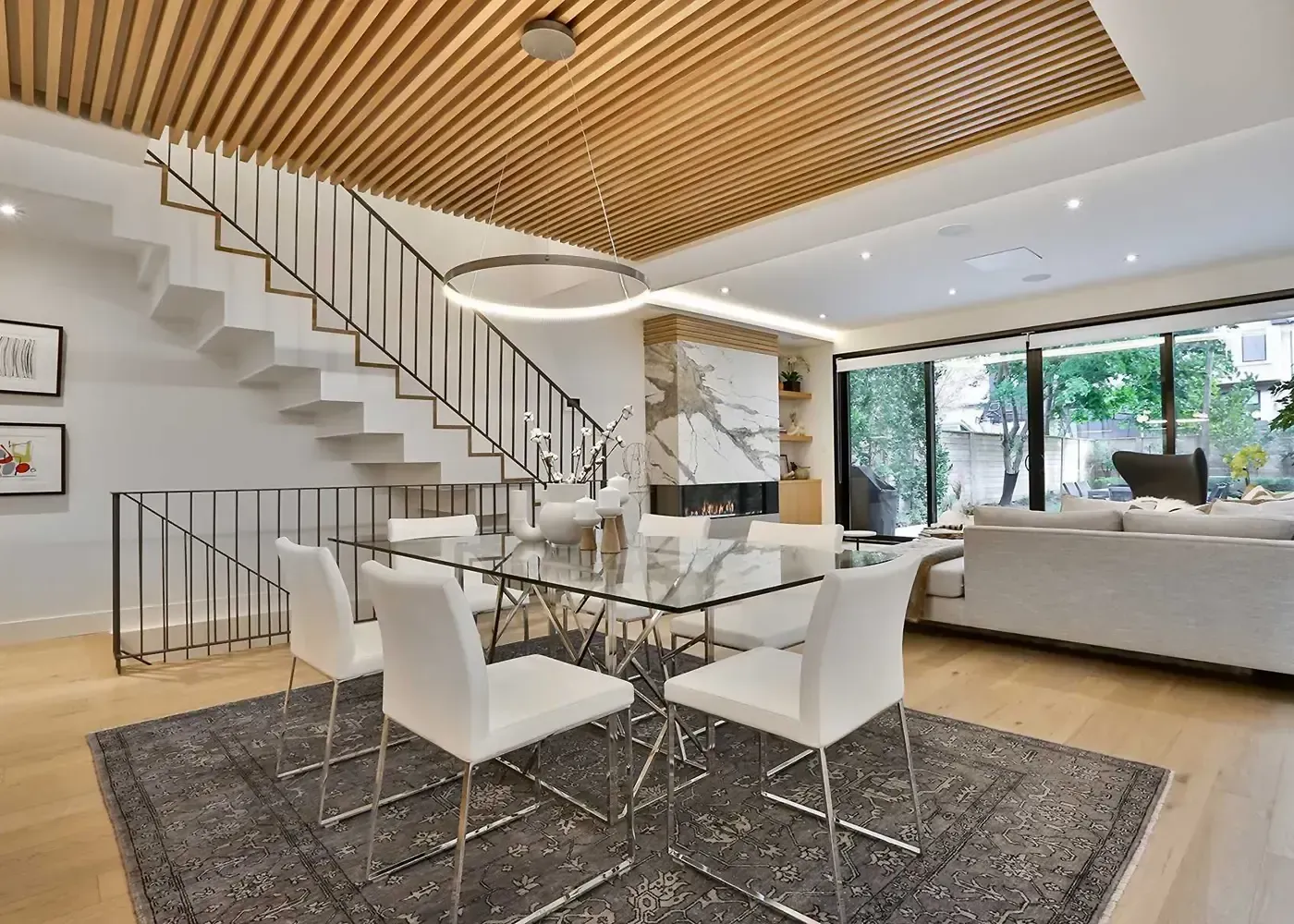 Modern dining room with glass table, white chairs, staircase, fireplace, and wood-slat ceiling.