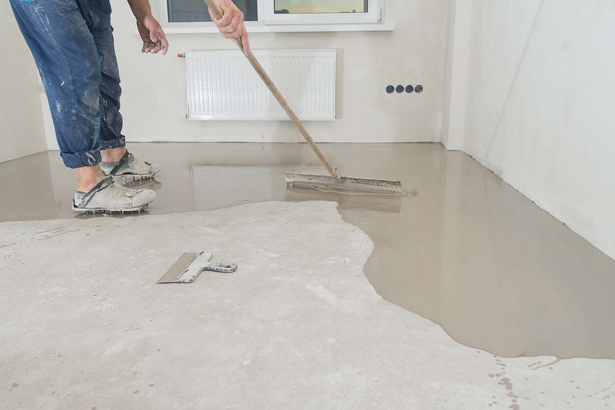 Person smoothing wet cement floor with a long-handled tool; room with white walls, window, and baseboard heater.