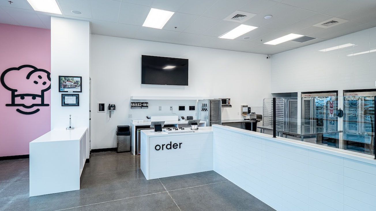 Interior of a bakery with a pink wall and white counter. Kitchen equipment visible.