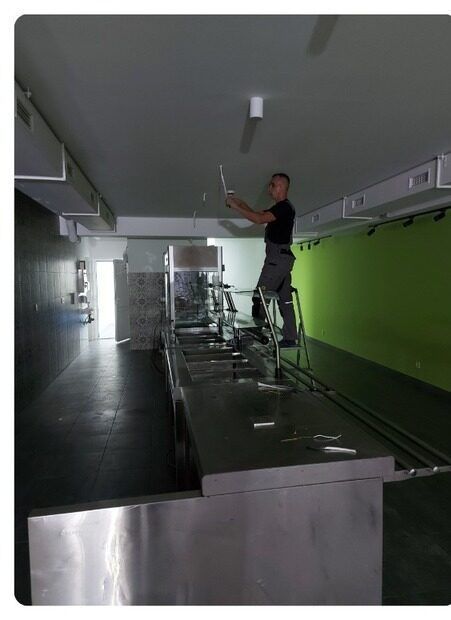 Man on a ladder installing lights in a commercial kitchen with stainless steel counters and green wall.